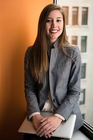 A young woman in a business suit sitting in front of a window, looking focused and professional.